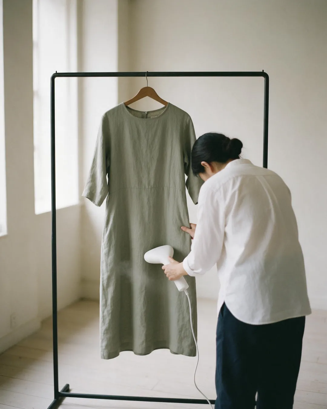 A woman in a white shirt and navy trousers, seen from behind with her head bowed, steams a sage-green linen midi dress hanging on a black clothing rail with a white handheld steamer in a daylit white room.