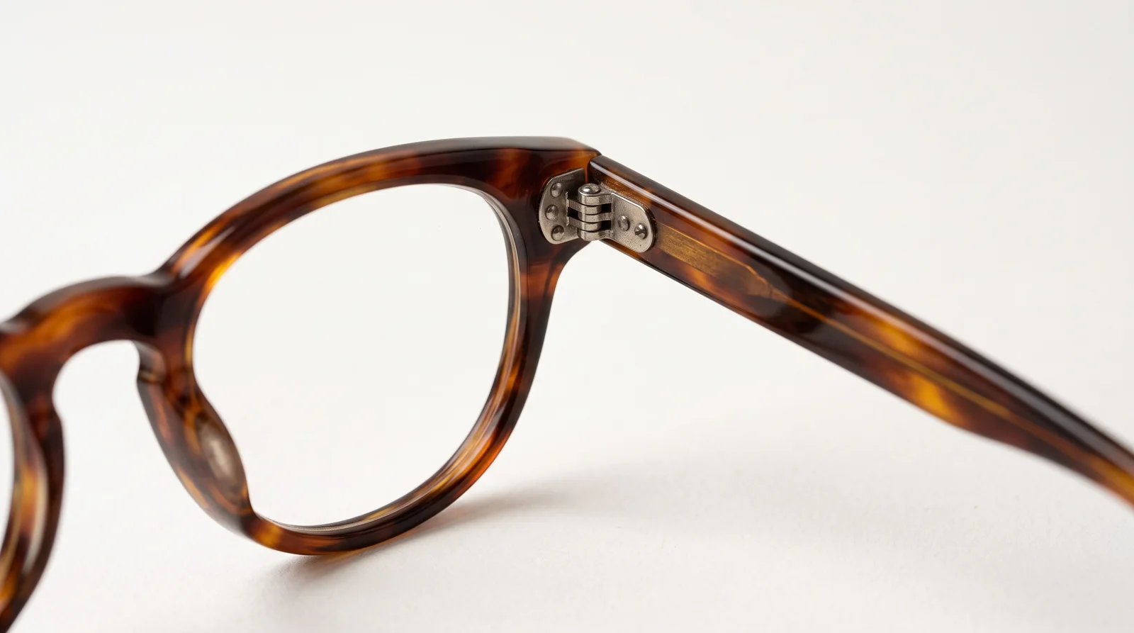 Close-up of a tortoiseshell acetate frame on white, focused on the metal hinge where the temple meets the front, with a clear lens and no glare.