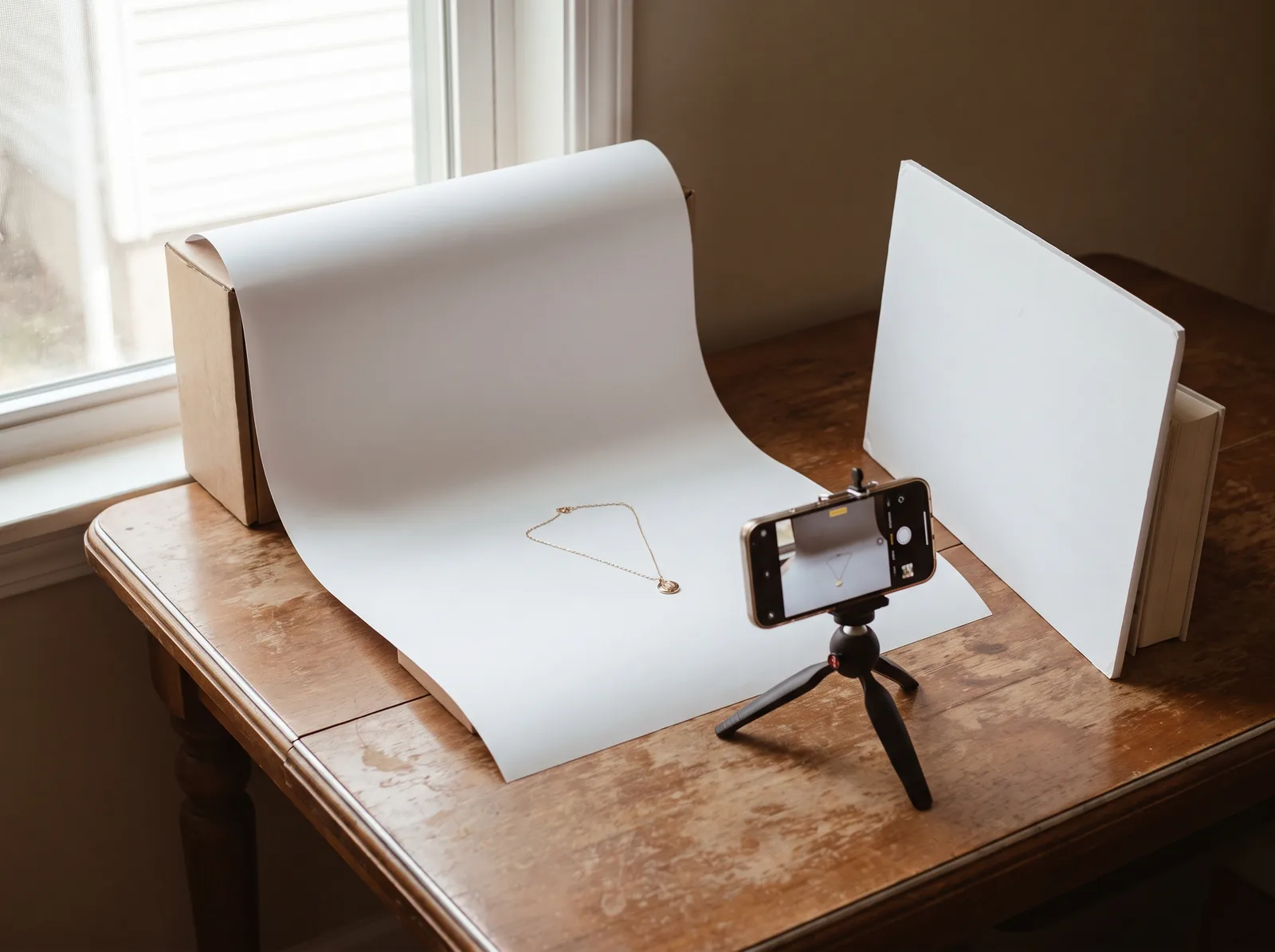 A home jewelry photography setup: a gold necklace on a white paper sweep by a window, a phone on a small tripod and a white card reflector