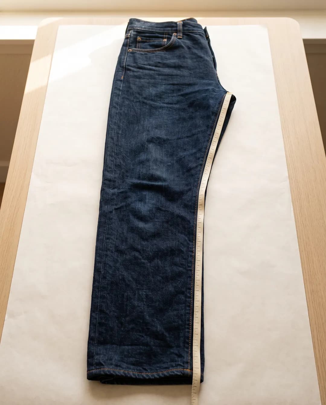 Dark-wash jeans folded in half lengthwise on white paper, with a cream tape measure laid along the inner leg seam from the crotch to the hem