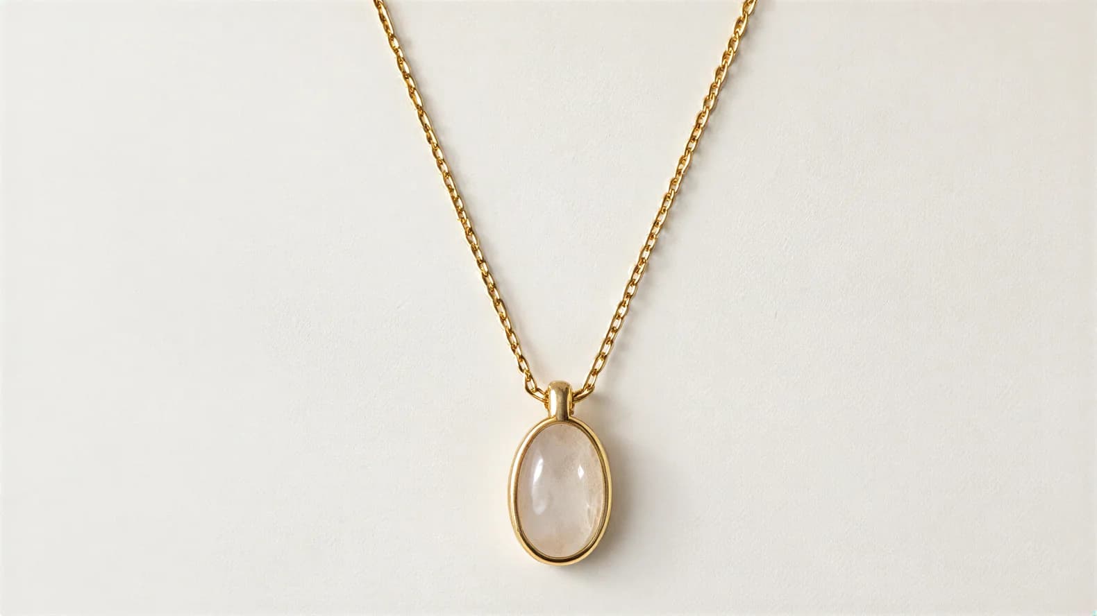 A gold chain necklace with an oval pale stone pendant on a plain white surface