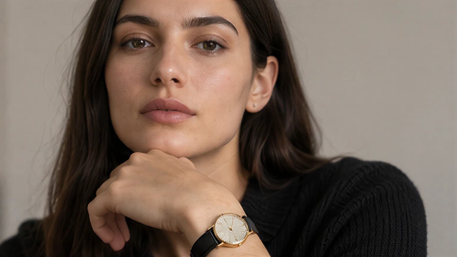 A model in a black knit sweater resting her chin on her hand, wearing the gold dress watch on a black leather strap