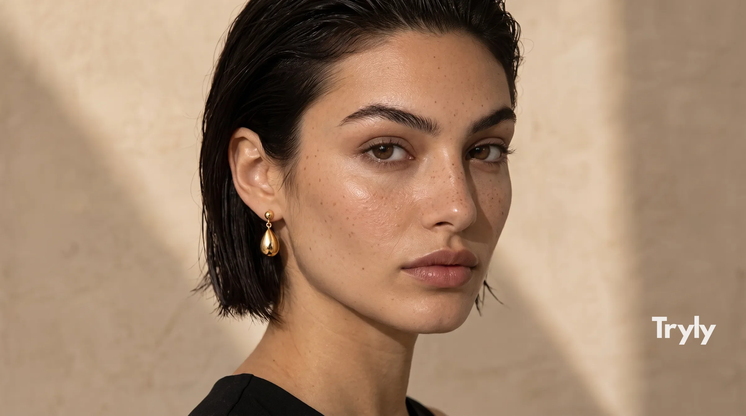 A woman with short dark hair wearing gold teardrop drop earrings, in warm window light