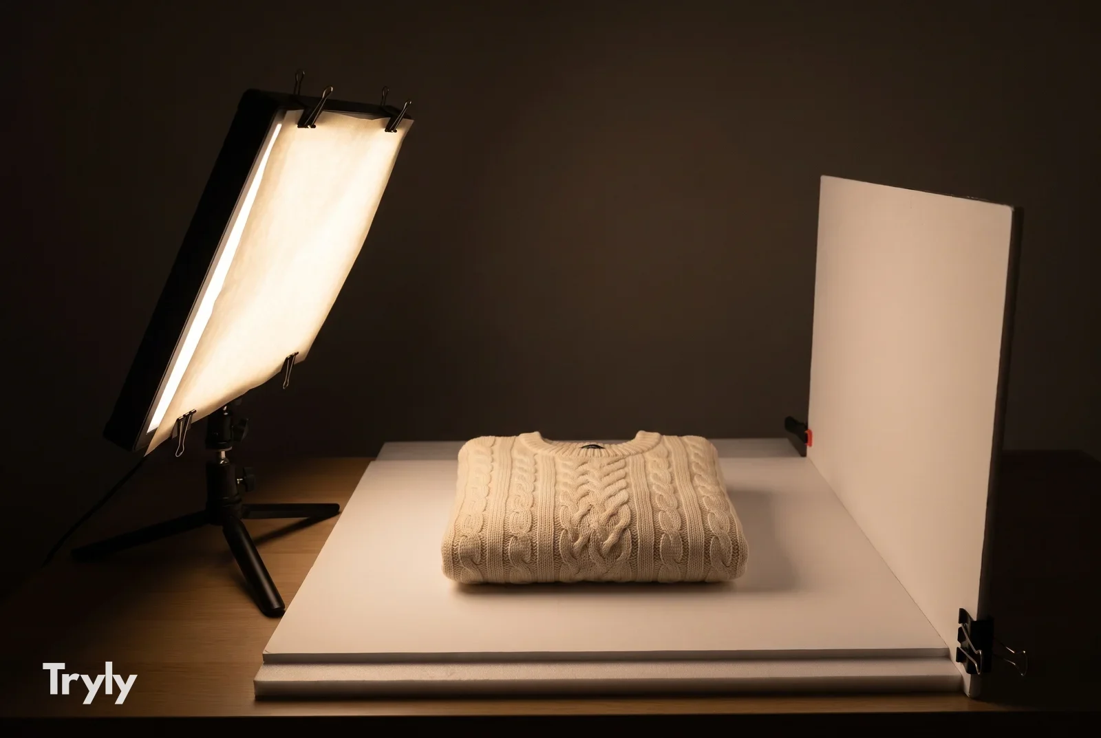 A folded cream cable-knit sweater on a white foam board in a dark room, lit from the left by one LED panel on a small tabletop tripod with baking parchment clipped over its face, and an upright white foam board on the right bouncing light back.