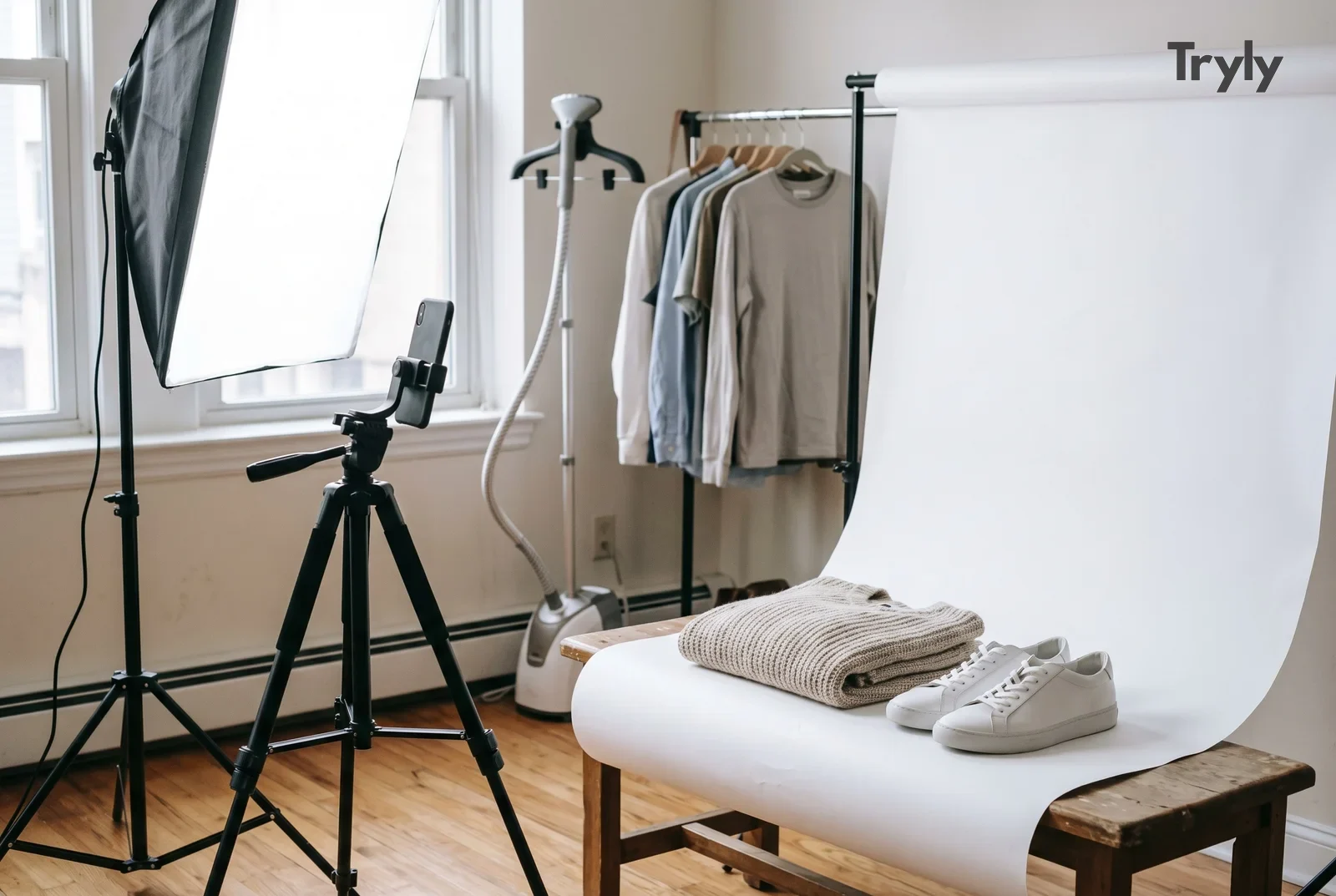 A home product-photo corner: a white paper sweep over a wooden bench with a folded knit sweater and white sneakers, a softbox by the window, a phone on a tripod, a steamer and a clothing rail.